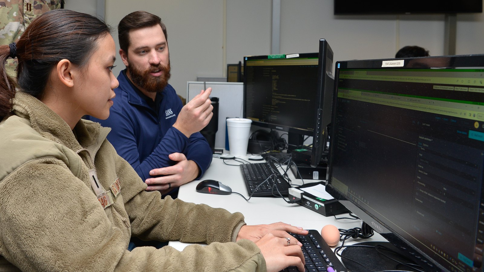 A military cyber operator and a civilian analyst review information on computer monitors.