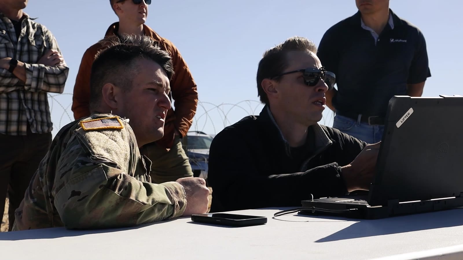 Military and civilian colleagues review a laptop together during an outdoor technology test.