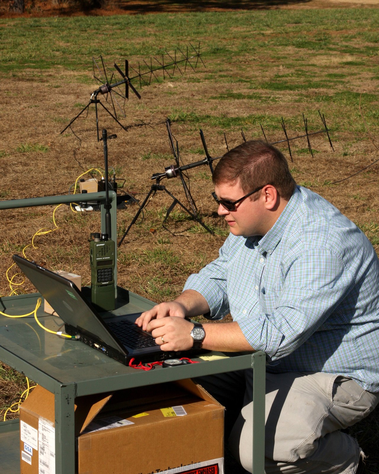 A field technician uses a laptop beside radio equipment and directional antennas outdoors.