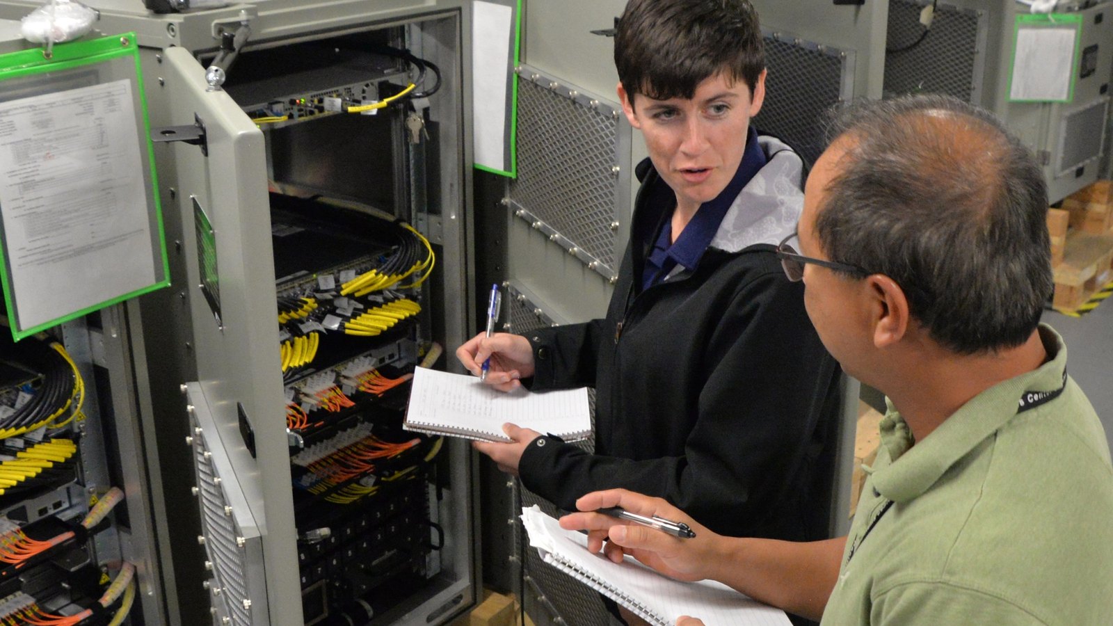 Two technicians review notes beside open network racks filled with connected equipment.
