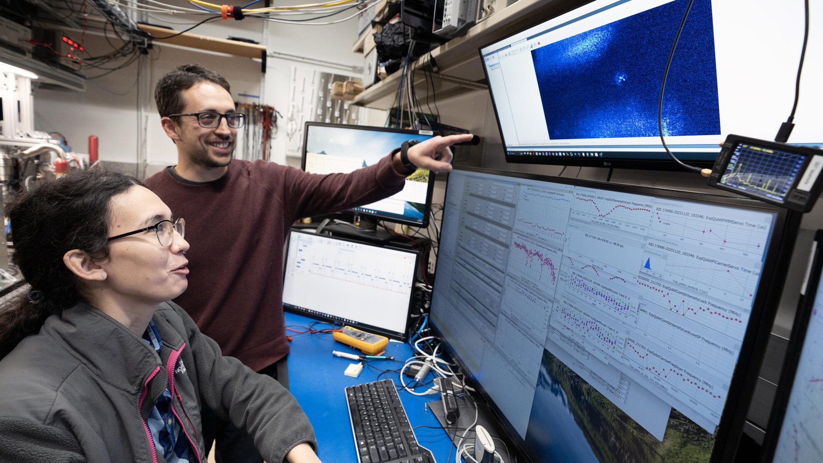Two researchers study experiment data across several monitors in an electronics laboratory.