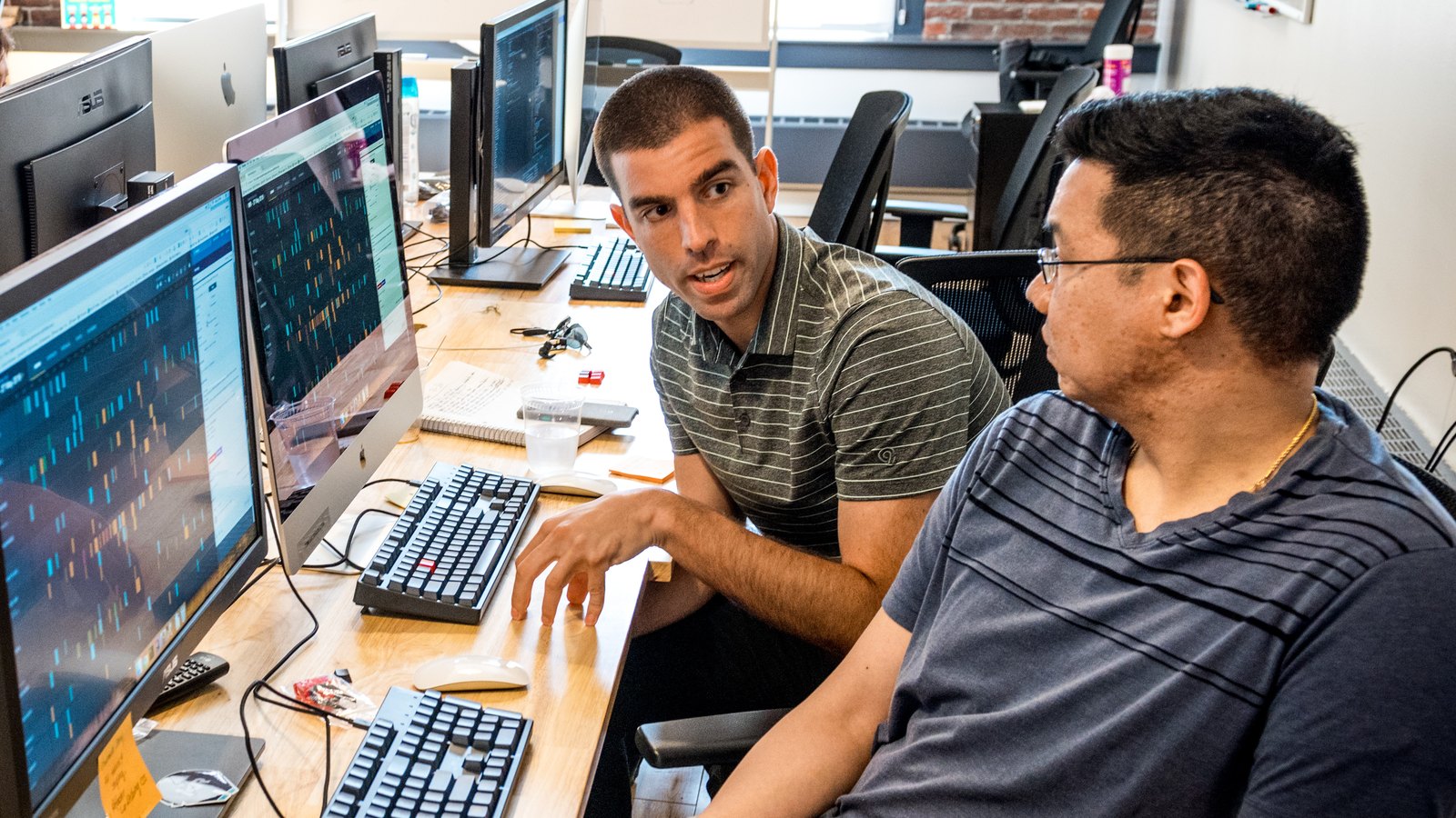 Software developers collaborate at shared computer workstations in a bright office.