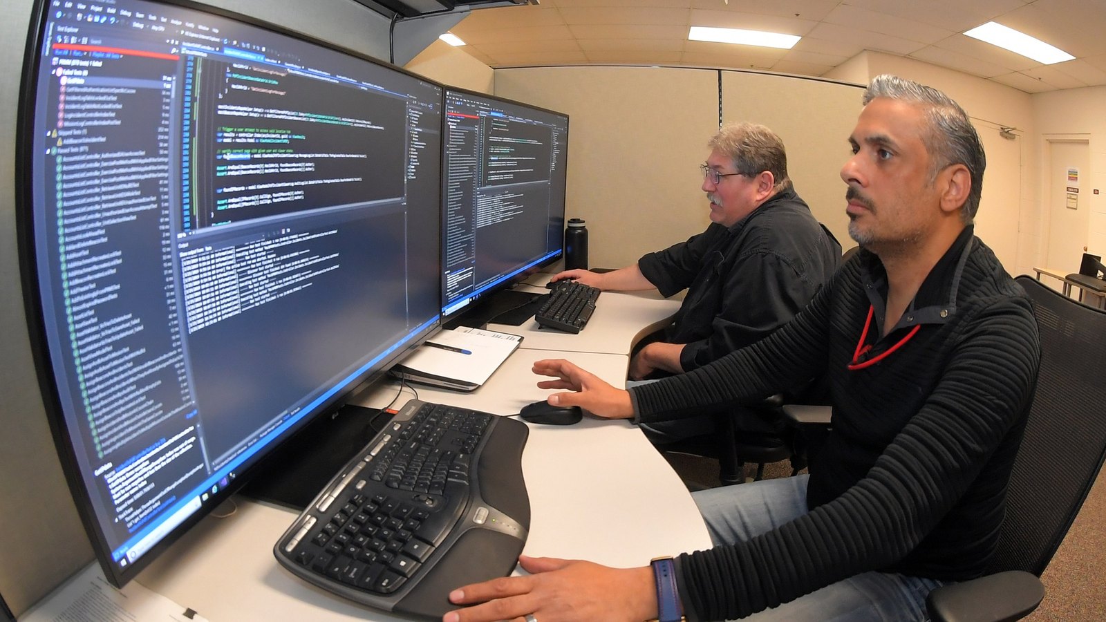 Two civilian software engineers work side by side at monitors displaying code.
